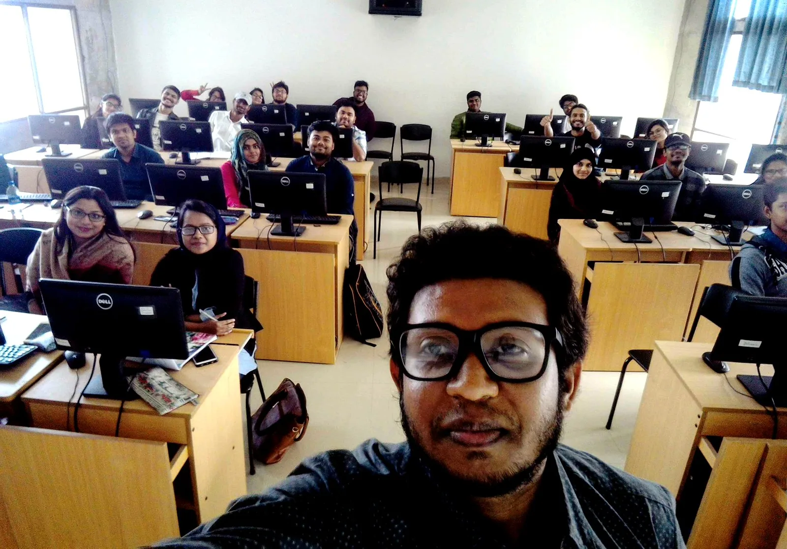 Sabbir Khan in a university computer lab at United International University, with around twenty students at desktop workstations behind him, several giving thumbs up.